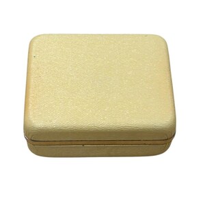 May include: A closed, square, cream-colored box with a textured surface. The box has a gold-colored trim around the edges. The box is likely used for storing jewelry or other small items.
