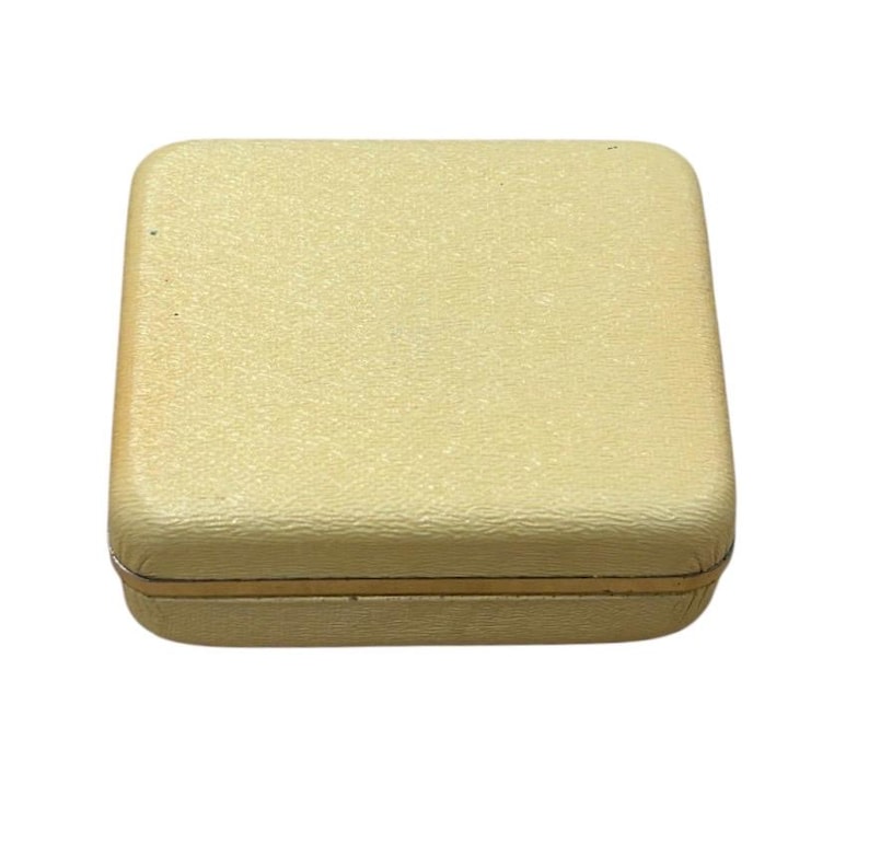 May include: A closed, square, cream-colored box with a textured surface. The box has a gold-colored trim around the edges. The box is likely used for storing jewelry or other small items.