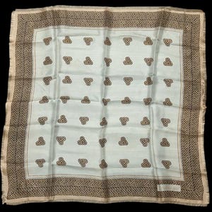 May include: A square, light blue scarf with a repeating brown knot pattern. The scarf has a brown patterned border and a fringed edge. The scarf is likely made of silk or a similar material.