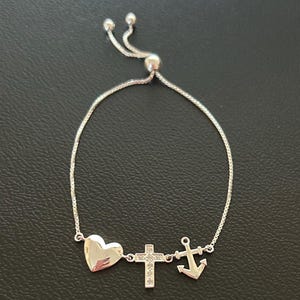 May include: A silver adjustable bracelet with a heart, cross, and anchor charm. The charms are all silver and have a shiny finish.