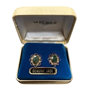May include: Open vintage jewelry box with a pair of gold-toned earrings. Each earring features a green oval jade stone surrounded by a decorative gold filigree design. The box is cream-colored with the text "14 KT. GOLD" and a label that reads "GENUINE JADE".