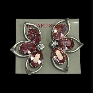 May include: A pair of silver tone earrings with pink and clear rhinestones in a flower shape. The earrings are on a white card with the text "Richard Serbin" printed on it.