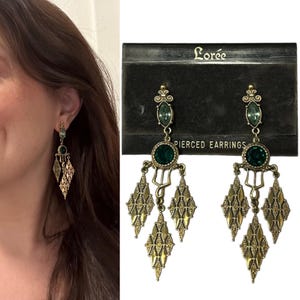 May include: A pair of gold tone dangle earrings with green crystal accents. The earrings are on a black card with the text "Lorée Pierced Earrings".