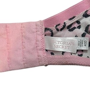 May include: Pink bra with a leopard print lining. The bra features a hook-and-eye closure and a tag that reads "VICTORIA'S SECRET" and size 40D. The bra is made of a soft, comfortable material.