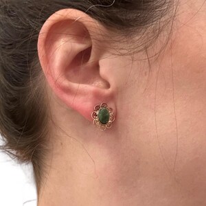 May include: A close-up of a gold-colored stud earring with a dark green oval stone. The earring has a floral filigree design around the stone. The earring is on a person's ear.
