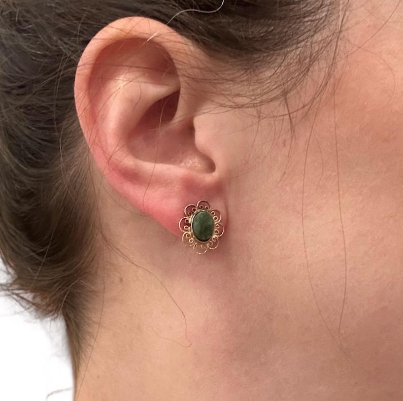 May include: A close-up of a gold-colored stud earring with a dark green oval stone. The earring has a floral filigree design around the stone. The earring is on a person's ear.