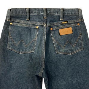 Op de afbeelding: Donkerblauwe denim jeans met klassieke vijf-zakken-stijl. Kenmerken zijn koperen klinknagels, een leren patch met het "Wrangler"-logo en kenmerkende "W"-stiksels op de achterzakken. Een tijdloze garderobe-klassieker.