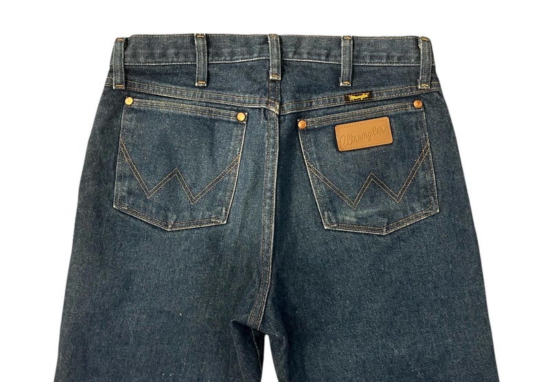 Op de afbeelding: Donkerblauwe denim jeans met klassieke vijf-zakken-stijl. Kenmerken zijn koperen klinknagels, een leren patch met het "Wrangler"-logo en kenmerkende "W"-stiksels op de achterzakken. Een tijdloze garderobe-klassieker.