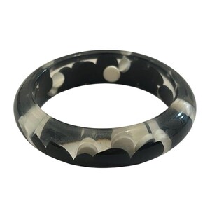 May include: A circular bangle bracelet made of a smooth, translucent material. The bracelet features a repeating pattern of solid black and opaque white circular shapes embedded within the clear, glossy resin structure of the band.