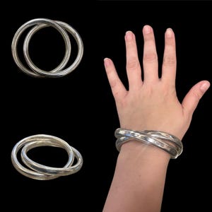Puede incluir: Un brazalete plateado con tres bandas redondeadas entrelazadas. La imagen muestra el brazalete en una muñeca y dos vistas adicionales sobre un fondo negro. El brazalete tiene un diseño simple y elegante.