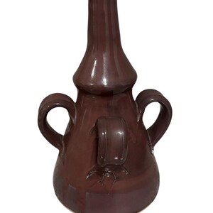 May include: A maroon ceramic vase with a bulbous base, a central section with a handle, and two side handles. The vase has a tall, slender neck and a glossy finish. The design includes a decorative element near the handle.
