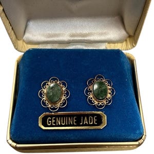 May include: A pair of oval green jade earrings set in gold filigree, displayed in a vintage gold box with a blue velvet interior. The box is labeled "14 KT. GOLD" and includes a tag that reads "GENUINE JADE".