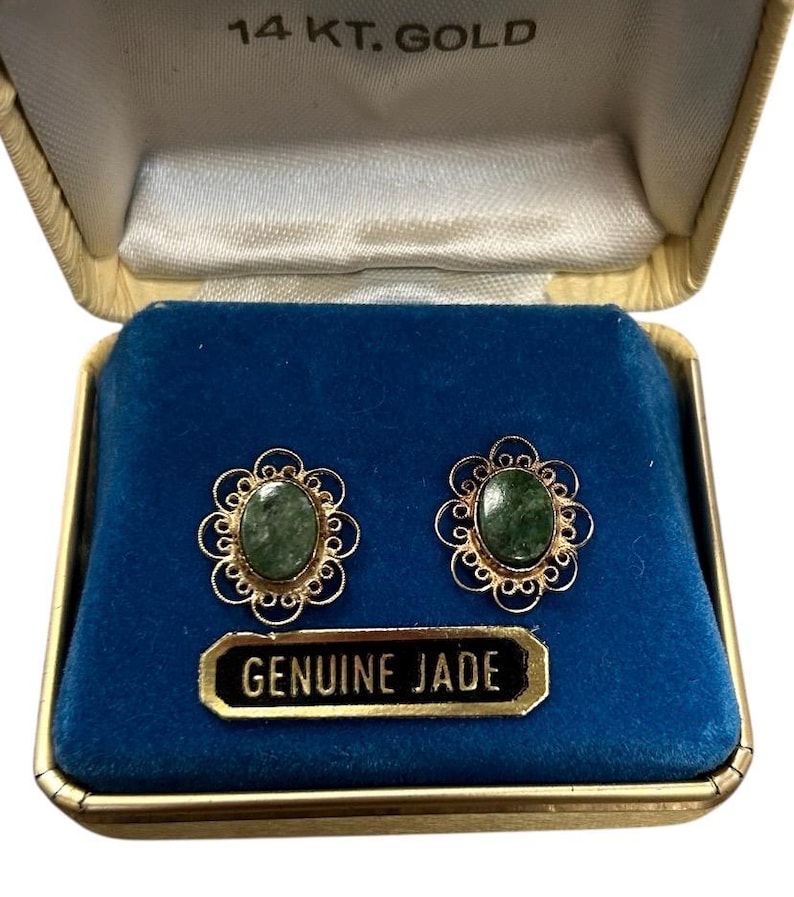 May include: A pair of oval green jade earrings set in gold filigree, displayed in a vintage gold box with a blue velvet interior. The box is labeled "14 KT. GOLD" and includes a tag that reads "GENUINE JADE".