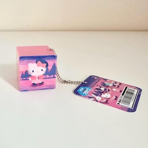 Puede incluir: Un llavero rosa en forma de cubo con un diseño de Hello Kitty. El diseño presenta a Hello Kitty con un atuendo rosa, sobre un paisaje de árboles y una puesta de sol. Una cadena plateada conecta el cubo a una etiqueta con texto japonés y un código de barras.
