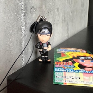 Può includere: Una piccola statuetta di Sai della serie anime Naruto, con un abito nero, guanti neri e una fascia. La statuetta è attaccata a un cordino nero. Sullo sfondo è visibile una colorata carta di Naruto.