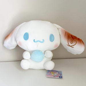 Puede incluir: Un peluche blanco de un personaje de dibujos animados con orejas grandes, ojos azules y una esfera azul claro. Las orejas tienen detalles marrones y un diseño de corazón. Se ve una pequeña tarjeta con personajes de dibujos animados.