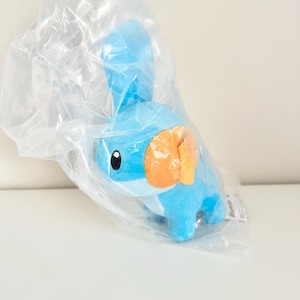 Op de afbeelding: Een blauw pluche speelgoed van een Mudkip-personage, met oranje wangvinnen, in een doorzichtige plastic zak. Het speelgoed heeft zwarte ogen en een witte buik. De pluche is een populair personage uit de Pokemon-franchise.