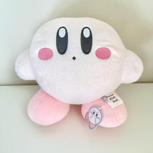 May include: A plush pink character with large black eyes, rosy cheeks, and small arms and feet. A tag attached to the foot reads "Sweet Pink Ball." The character is a popular video game icon.