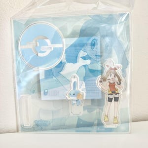 Op de afbeelding: Een doorzichtige plastic verpakking met een lichtblauwe standaard met een wit en lichtblauw ontwerp, een klein blauw cartoonfiguur en een cartoonfiguur met een rode top, zwarte shorts en gele schoenen. De achtergrond heeft een lichtblauwe illustratie.