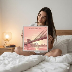 May include: A framed print featuring an illustration of the Brooklyn Bridge in New York City. The artwork depicts the bridge with a cityscape and a sunset in the background. The text "BROOKLYN BRIDGE - NYC" is at the top. The color palette includes shades of pink, orange, and gray.