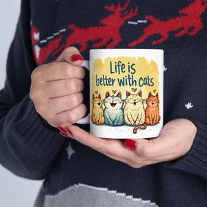 May include: White ceramic mug with the text "Life is better with cats" and four cartoon cats in different colours. The cats are sitting together with heart accents. The mug is held by a person.