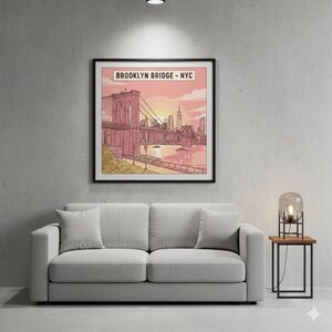 May include: A framed art print of the Brooklyn Bridge in New York City. The artwork features a pink and orange sunset over the bridge and city skyline. The print is displayed above a gray sofa with two pillows.