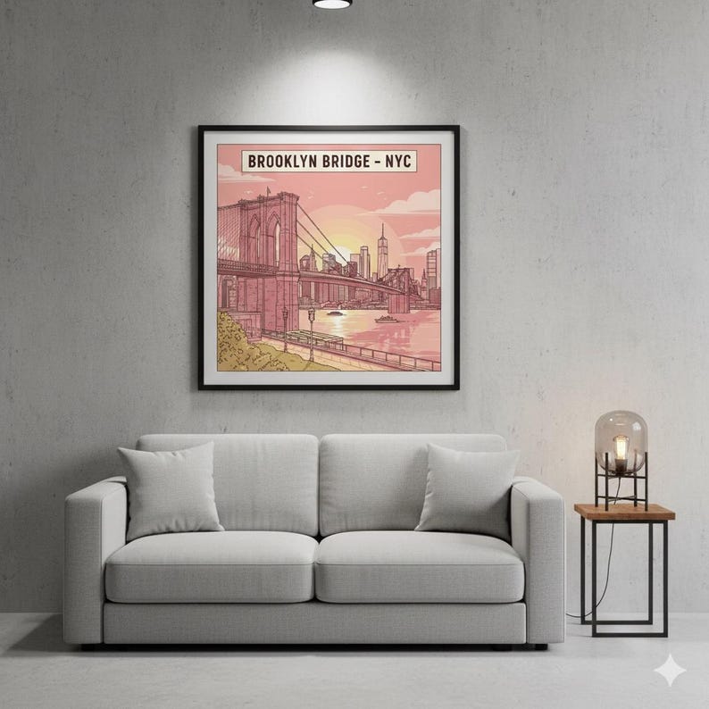 May include: A framed art print of the Brooklyn Bridge in New York City. The artwork features a pink and orange sunset over the bridge and city skyline. The print is displayed above a gray sofa with two pillows.