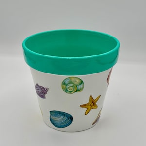 May include: White ceramic flower pot with a teal rim and seashell illustrations. The pot features a variety of colorful seashells and a yellow starfish design. The pot is designed for indoor or outdoor use.