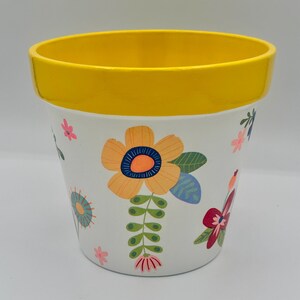 May include: White ceramic flower pot with a yellow rim, decorated with colorful floral designs. The pot features large orange and blue flowers, along with smaller pink and blue floral accents. A decorative item for home or garden.