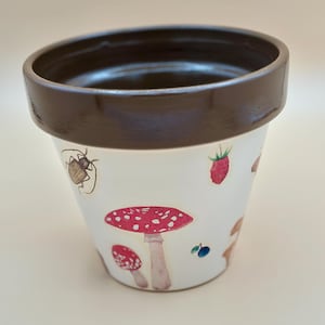 May include: White ceramic flower pot with a brown rim, decorated with illustrations of red mushrooms, strawberries, and insects. The pot is designed for planting flowers or herbs.