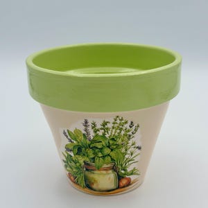 May include: A small, beige ceramic flower pot with a light green rim. The pot features a colorful illustration of various herbs and a jar. The design includes green, purple, and brown tones, perfect for a kitchen or garden.