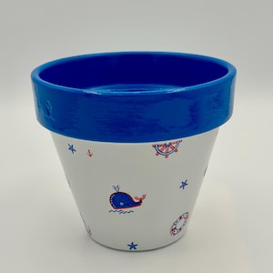 May include: A white ceramic flower pot with a blue rim and nautical-themed illustrations. The pot features a blue whale, red and blue anchors, stars, and life preservers. The pot is approximately 5 inches tall.