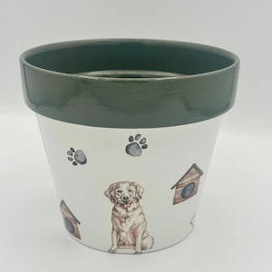 May include: White ceramic flower pot with a dark green rim. The pot features a watercolor illustration of a golden retriever, paw prints, and dog houses. A decorative item for dog lovers.