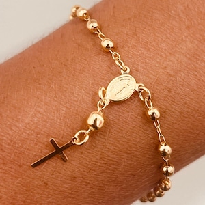 May include: Gold-colored rosary bracelet featuring a cross pendant and a central medallion. The bracelet is composed of small, round beads and delicate chain links. The design is simple and elegant.