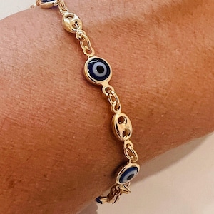 May include: Gold-coloured bracelet with blue and white evil eye charms. The bracelet has a delicate chain design with oval links and is worn on a wrist. The evil eye charms are round with a blue iris and black pupil.