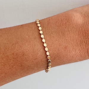 May include: A gold bracelet with a chain of small, flat, circular links. The bracelet is worn on a wrist, highlighting its simple design. The warm gold colour contrasts with the skin tone, highlighting the jewellery's subtle shine. The bracelet's length is approximately 18 cm.