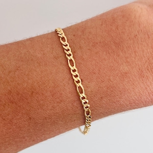 May include: A gold-colored Figaro chain bracelet. The bracelet features a pattern of three small circular links followed by one elongated oval link. The bracelet is worn on a wrist.