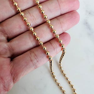 May include: A gold-colored ball chain necklace. The necklace is composed of small, evenly spaced, spherical beads. The chain is held against a light-colored background, highlighting the necklace's shiny, metallic appearance. The necklace is a jewelry item.