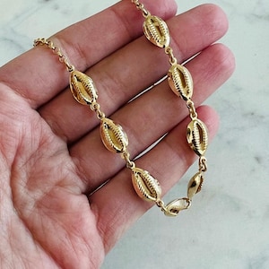 May include: A gold necklace with a series of seashell charms. The charms are oval-shaped with detailed ridges, and the chain is delicate. The necklace is displayed against a light background.
