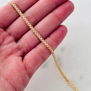 Op de afbeelding: Een delicate goudkleurige ketting met een geweven ontwerp. De ketting wordt in de hand van een persoon gehouden, waarbij het ingewikkelde patroon en de subtiele glans worden getoond. De ketting is ongeveer 50 cm lang.