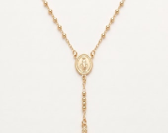 May include: A gold-toned rosary necklace featuring a delicate chain with small round beads. The center of the necklace displays an oval medal depicting a figure, and it terminates in a detailed crucifix pendant hanging from the bottom section.