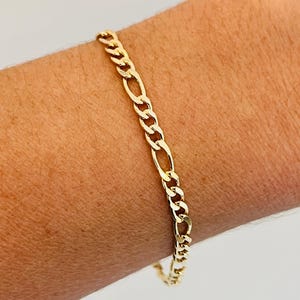 May include: A gold-coloured Figaro chain bracelet. The bracelet features a pattern of three small circular links followed by one elongated oval link. The bracelet is worn on a wrist.