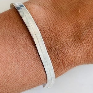 Puede incluir: Una pulsera de plata con diseño de espiga. La pulsera es plana y flexible, con una serie de pequeños eslabones entrelazados que crean una superficie texturizada. La pulsera se lleva en la muñeca.