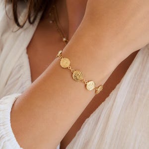 Può includere: Un bracciale a catena color oro caratterizzato da una serie di piccoli medaglioni circolari. Ogni medaglione presenta un dettagliato design in rilievo sulla superficie. Il bracciale è realizzato con delicati anelli metallici che collegano i ciondoli.