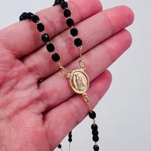 May include: A black beaded rosary with a gold-coloured medallion featuring a religious figure. The rosary is held in a hand, showcasing the details of the beads and the medallion's design. The beads appear to be faceted, reflecting light.