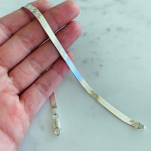Peut inclure: Un bracelet chaîne en argent en forme d'arête de poisson avec un fermoir mousqueton. Le bracelet est plat et souple, avec une surface texturée qui capte la lumière. Le bracelet est présenté sur un fond clair.