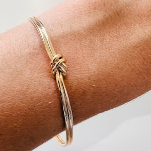 Pode incluir: Pulseira dourada com design de nó. A pulseira tem um acabamento liso e polido e um estilo simples e elegante. A pulseira é usada no pulso.