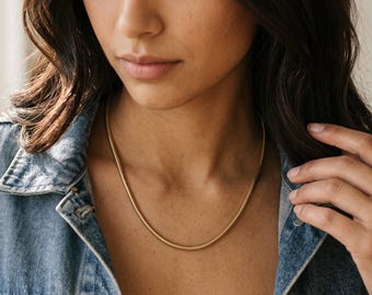 Könnte beinhalten: Eine goldfarbene Schlangenkette mit einem glatten, abgerundeten Profil und polierter Oberfläche. Die Kette liegt am Hals an und zeichnet sich durch ein durchgehendes, flexibles Glieddesign aus, das für ein elegantes, gleichmäßiges Erscheinungsbild sorgt.