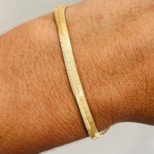 Puede incluir: Una pulsera dorada plana con diseño de espiga. La pulsera, que se lleva en la muñeca, tiene una superficie lisa y reflectante. El diseño es una serie de eslabones angulados e interconectados. La pulsera mide aproximadamente 0,5 cm de ancho.
