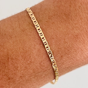 May include: A gold-colored chain bracelet with a flat, rectangular link design. The bracelet is worn on a person's wrist, showcasing its shiny, metallic appearance. The bracelet's links are evenly spaced, creating a continuous pattern.